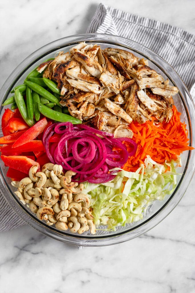 Large bowl with sliced chicken, shredded carrots, sliced cabbage, toasted cashews, pickled onions, sliced bell pepper, and snap peas.