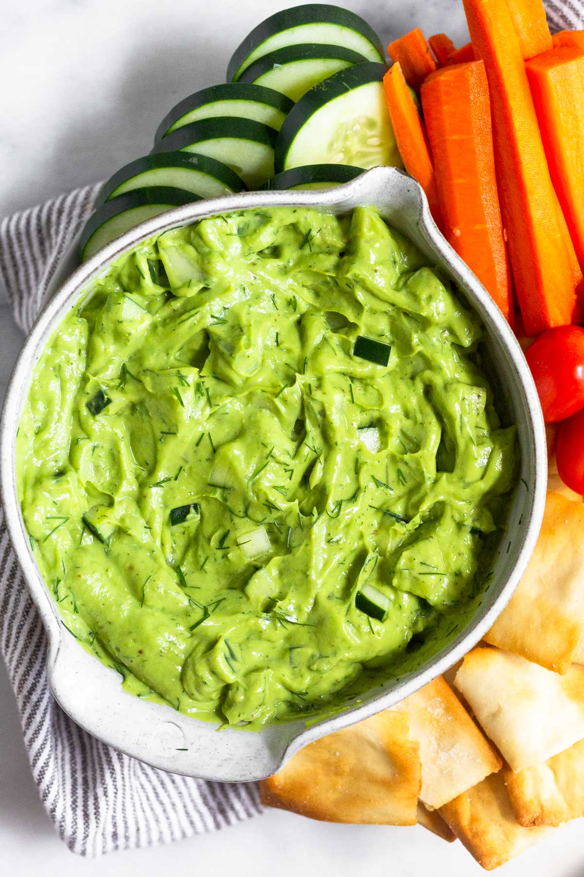 Avocado tzatziki dip in a bowl. Around it is pita chips, cherry tomatoes, carrot sticks, and sliced cucumber.