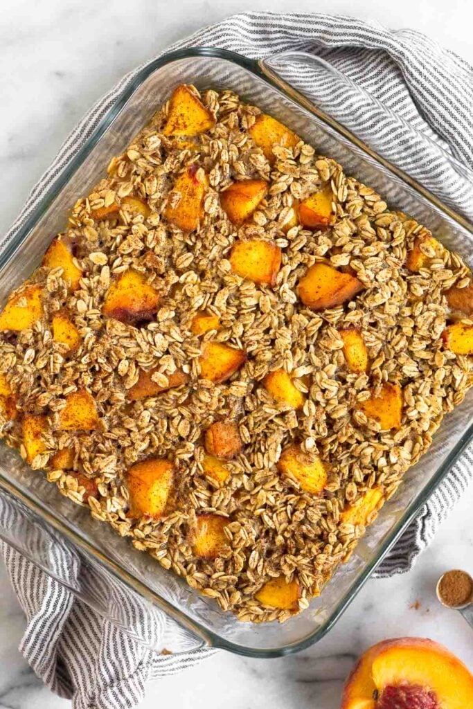 Peach baked oatmeal in a glass dish. It is sitting on a kitchen towel with half a peach next to it.