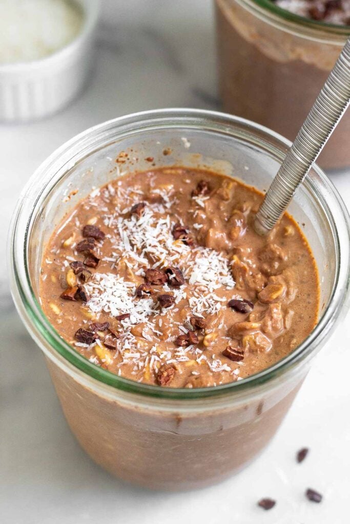 Jar of chocolate coconut overnight oats topped with cacao nibs and shredded coconut. There is spoon in the jar and behind it is another jar and a bowl of coconut.