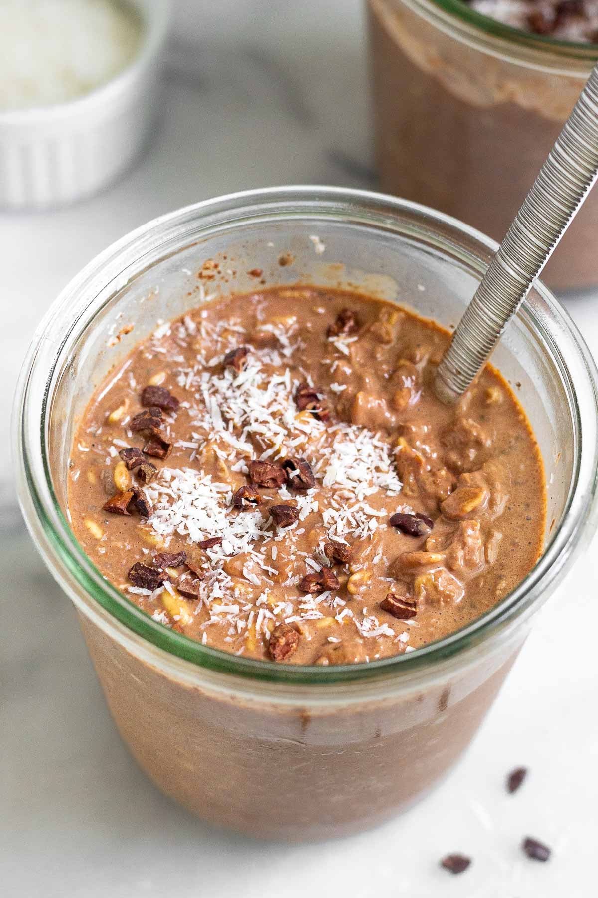 Jar of chocolate coconut overnight oats topped with cacao nibs and shredded coconut. There is spoon in the jar and behind it is another jar and a bowl of coconut.