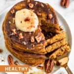 Overhead view of a stack of pumpkin pancakes with protein topped with coconut butter, chopped pecans, and maple syrup. A slices is missing from the stack and is on a fork that is laying on the plate.