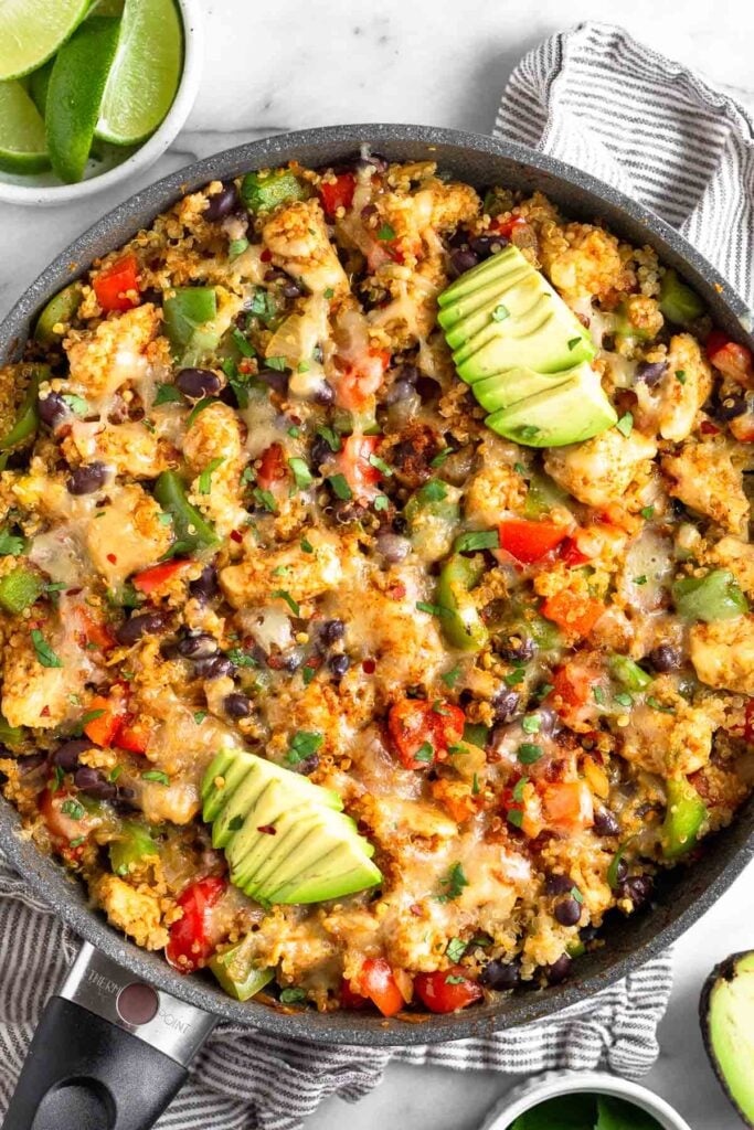 Chicken quinoa skillet in a large pan with veggies and topped with melted cheese topped with avocado. Around the pan is an half of avocado and a bowl of sliced lime.