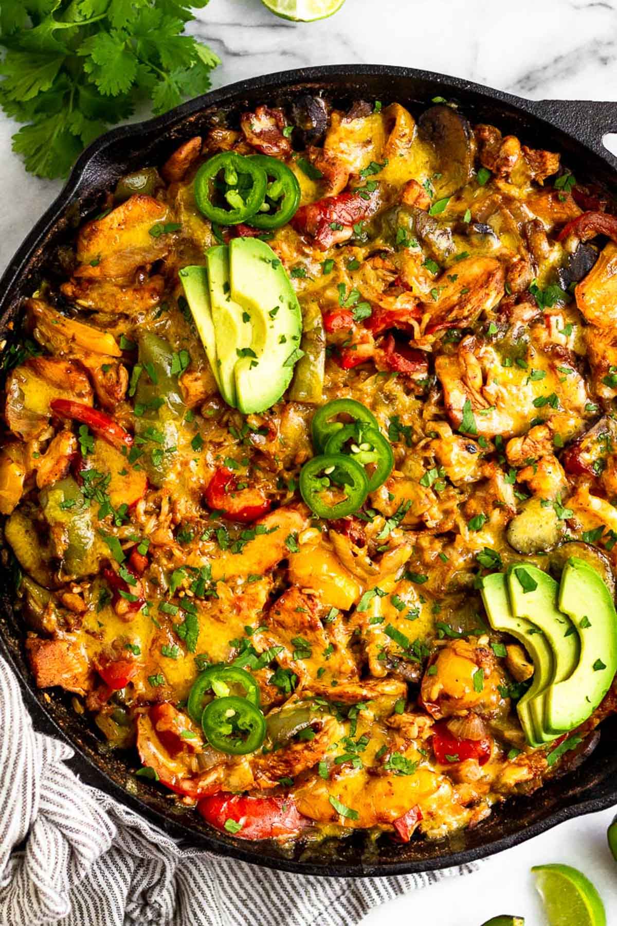 Healthy casserole recipe with chicken, rice, veggies, and cheese in a large cast iron. It is topped with avocado, jalapeños, and cilantro.