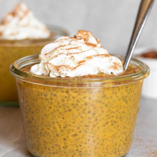 Jar of pumpkin chia pudding topped with whipped cream and cinnamon. A spoon is coming out of the jar and another jar is behind it.