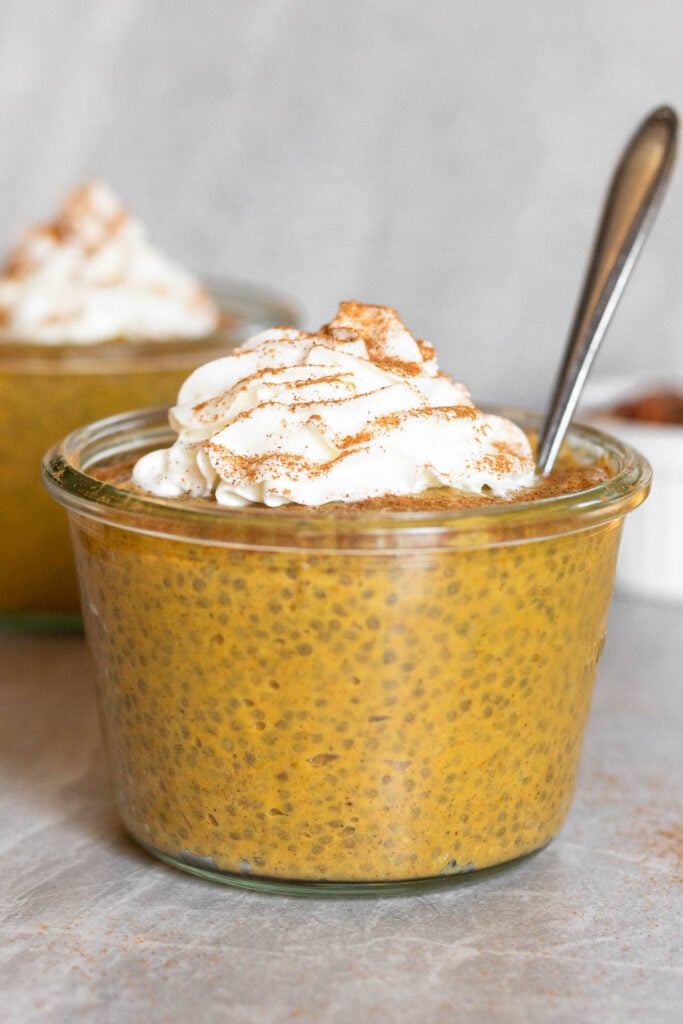 Jar of pumpkin chia pudding topped with whipped cream and cinnamon. A spoon is coming out of the jar and another jar is behind it.