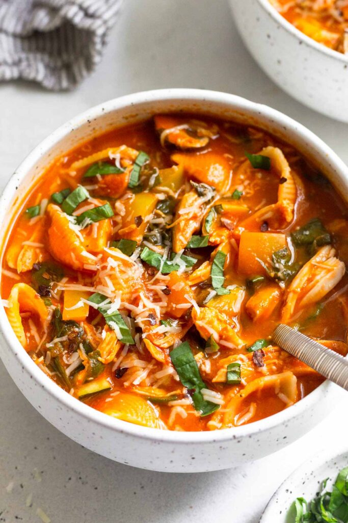 A bowl of chicken tomato soup topped with fresh herbs and cheese. A spoon is coming out of the bowl with a kitchen towel and another bowl behind it.