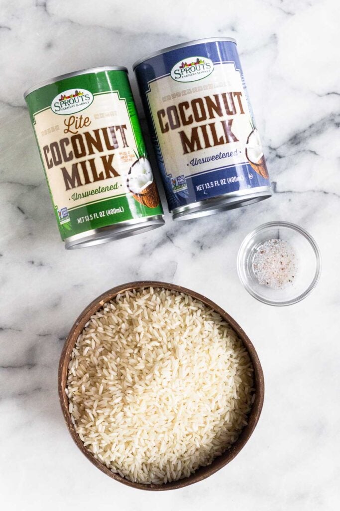 A can of lite coconut milk, a can of full fat coconut milk, a small bowl of salt, and a bowl of uncooked white rice on a marble countertop.