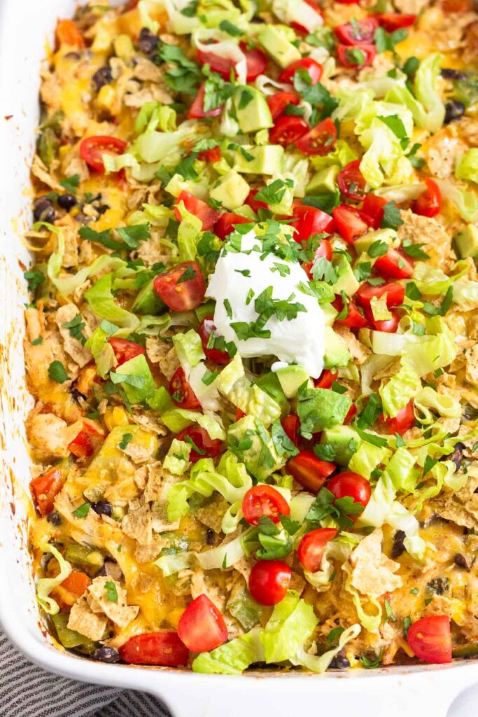 Chicken taco casserole in a large baking dish topped with shredded lettuce, diced tomatoes, diced avocado, sour cream, and cilantro.