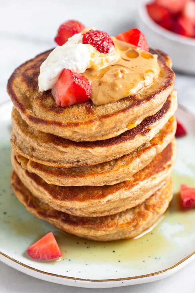 Close up of protein powder pancakes on a plate topped with yogurt, peanut butter, strawberries, and maple syrup.