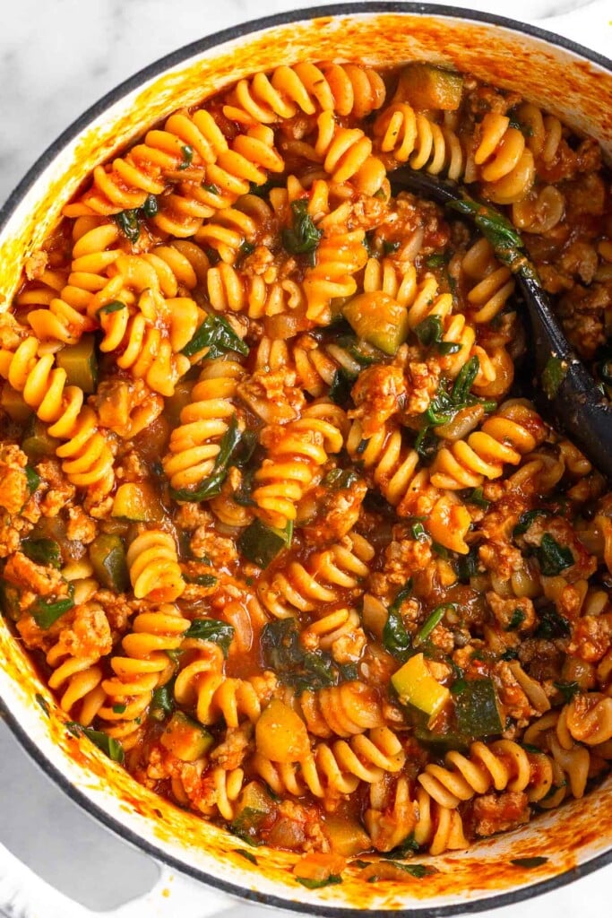 Large white dutch oven filled with ground turkey pasta with veggies. A spoon is also in the pot.