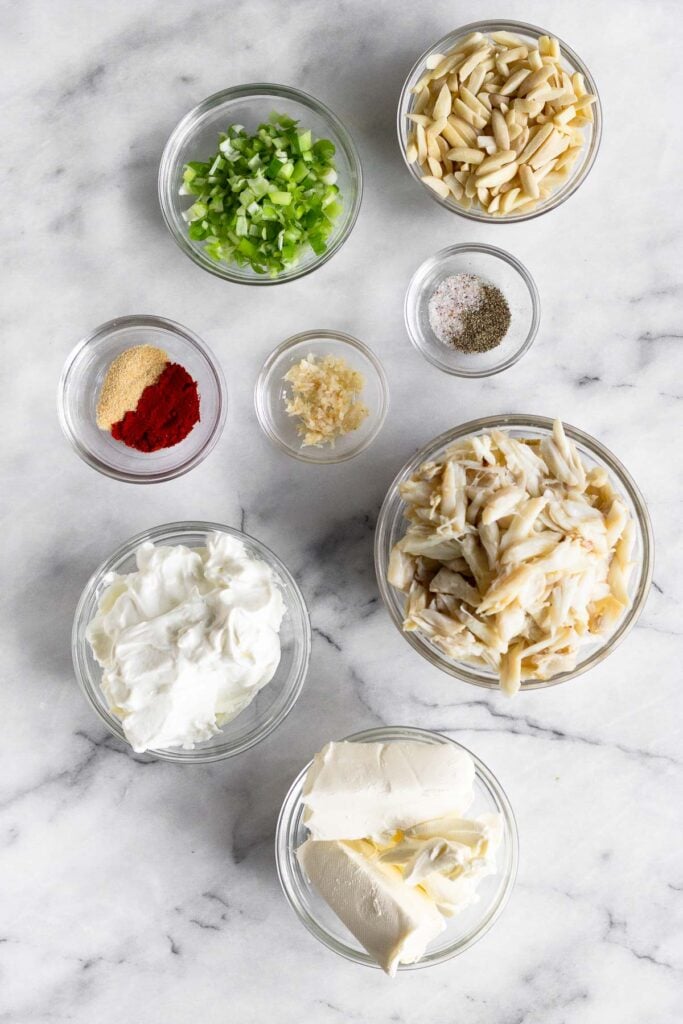 White counter top with a bowl of minced green onions, a bowl of slivered almonds, a bowl of salt and pepper, a bowl of lump crab meat, a bowl of cream cheese, a bowl of greek yogurt, a bowl of horseradish, and a bowl of spices.
