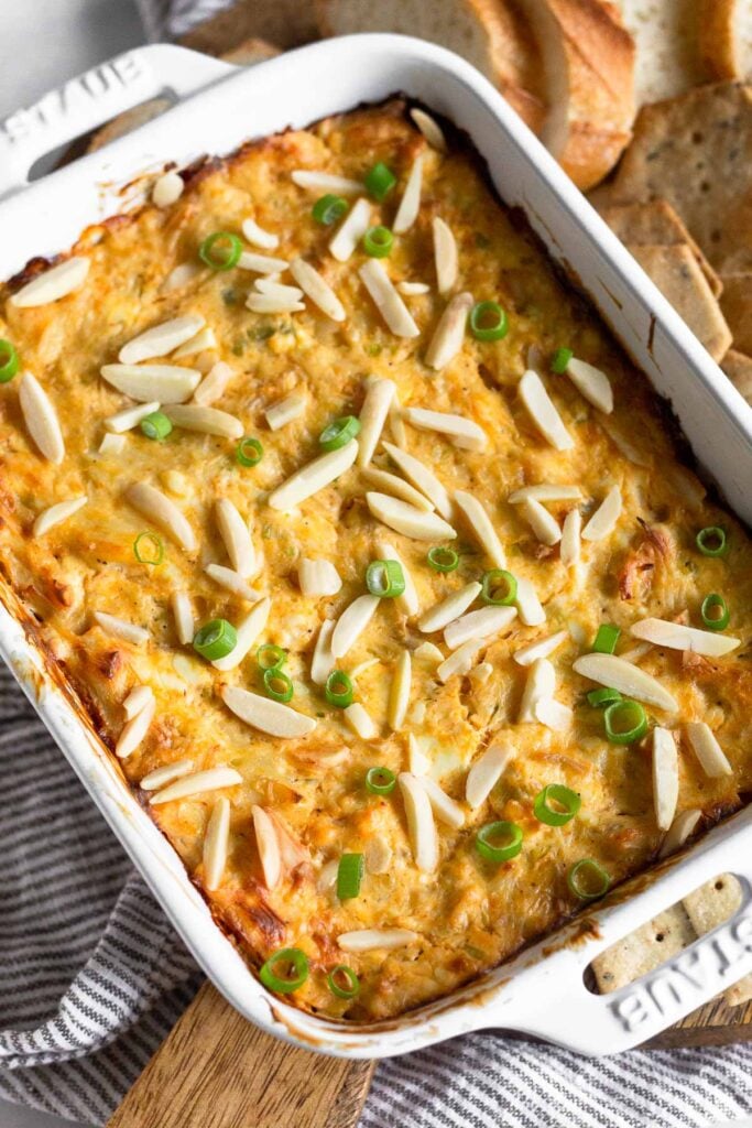 Baked crab dip in a white dish topped with slivered almonds and sliced green onions. Around it is a kitchen towel and some bread and crackers.