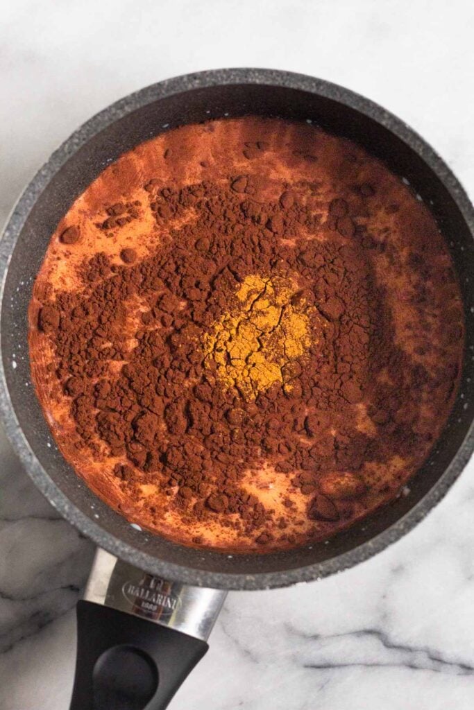 Saucepan filled with milk topped with cocoa powder and cinnamon before it is whisked together.
