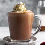 A glass mug filled with protein hot chocolate topped with whipped cream and cinnamon. The mug is on a small saucer and behind it is a jar of milk.