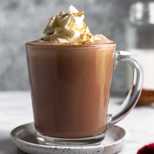 A glass mug filled with protein hot chocolate topped with whipped cream and cinnamon. The mug is on a small saucer and behind it is a jar of milk.