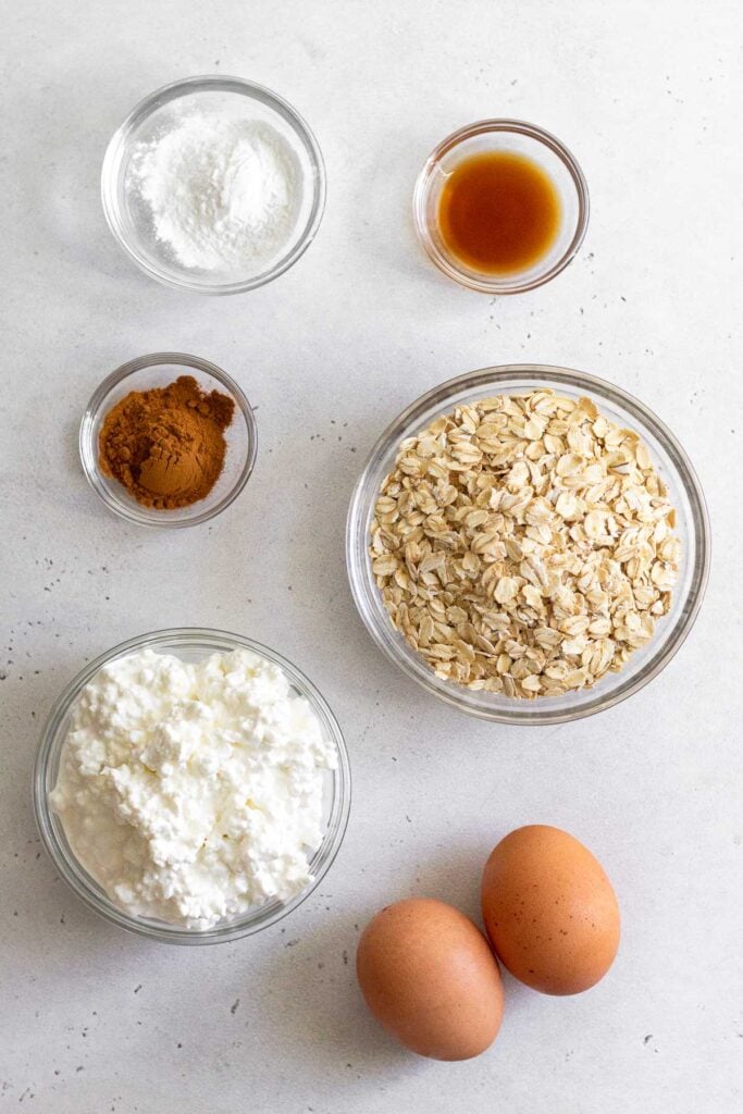 A small bowl of vanilla extract, a bowl of rolled oats, two large brown eggs, a bowl of cottage cheese, a small bowl of cinnamon, and a small bowl of baking powder on a white concrete counter.