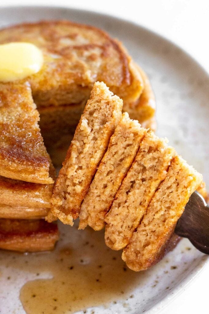 Flourless cottage cheese pancakes stacked on a plate with a section cut out of them on a fork. They are topped with some butter and maple syrup.