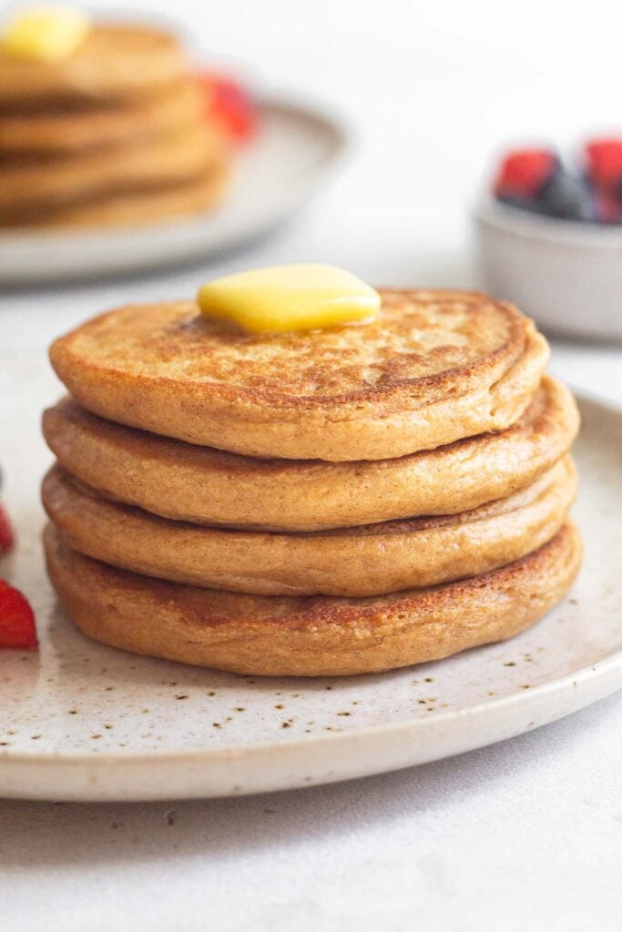 A stack of cottage cheese protein pancakes topped with butter on a plate. Behind the plate is a bowl of berries and another plate of a stack with pancakes.
