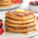 A plate of four cottage cheese pancakes topped with butter and syrup. There are some fresh berries on the plate. Behind the plate is a bowl of more berries and another stack of pancakes.