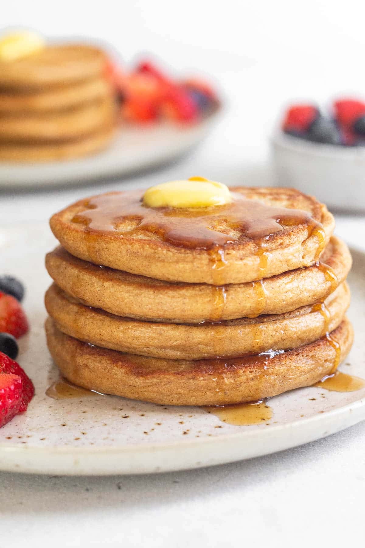 A plate of four cottage cheese pancakes topped with butter and syrup. There are some fresh berries on the plate. Behind the plate is a bowl of more berries and another stack of pancakes.