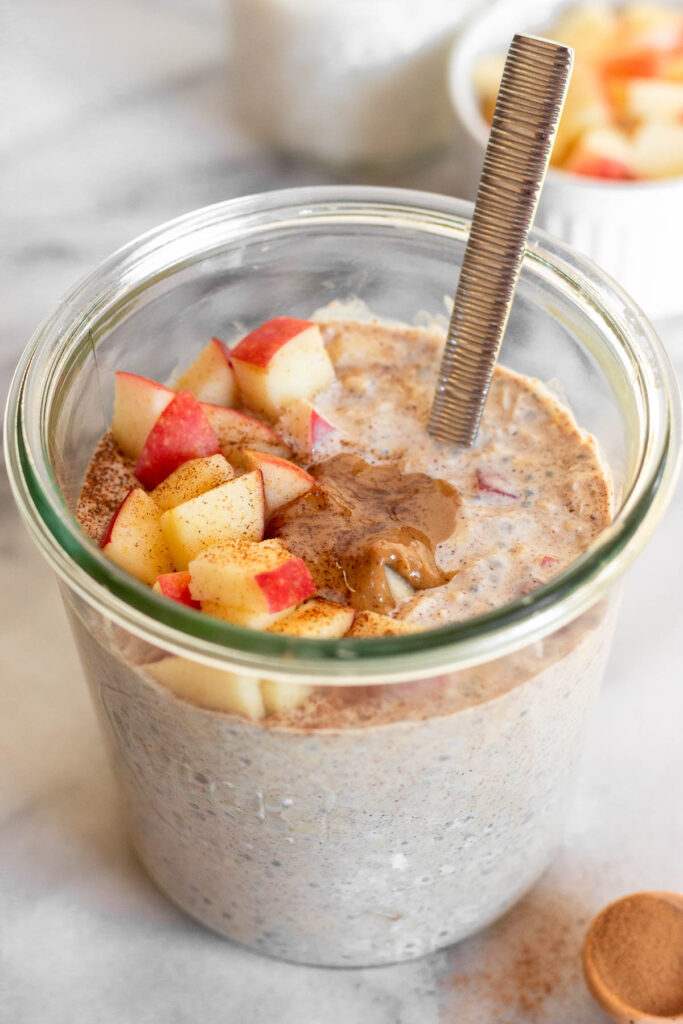 Overnight oats in a jar topped with apple, peanut butter, and cinnamon with a spoon coming out of the jar.