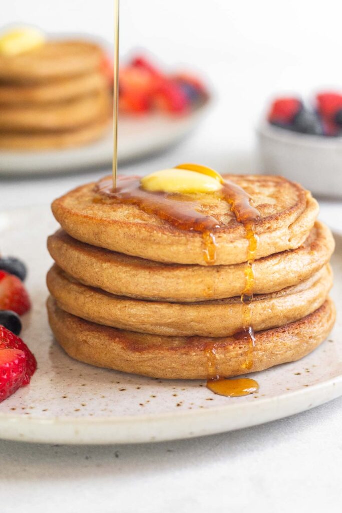 Cottage cheese pancakes on a plate topped with butter and maple syrup being poured on it. Behind it is another plate with pancakes and fresh fruit.