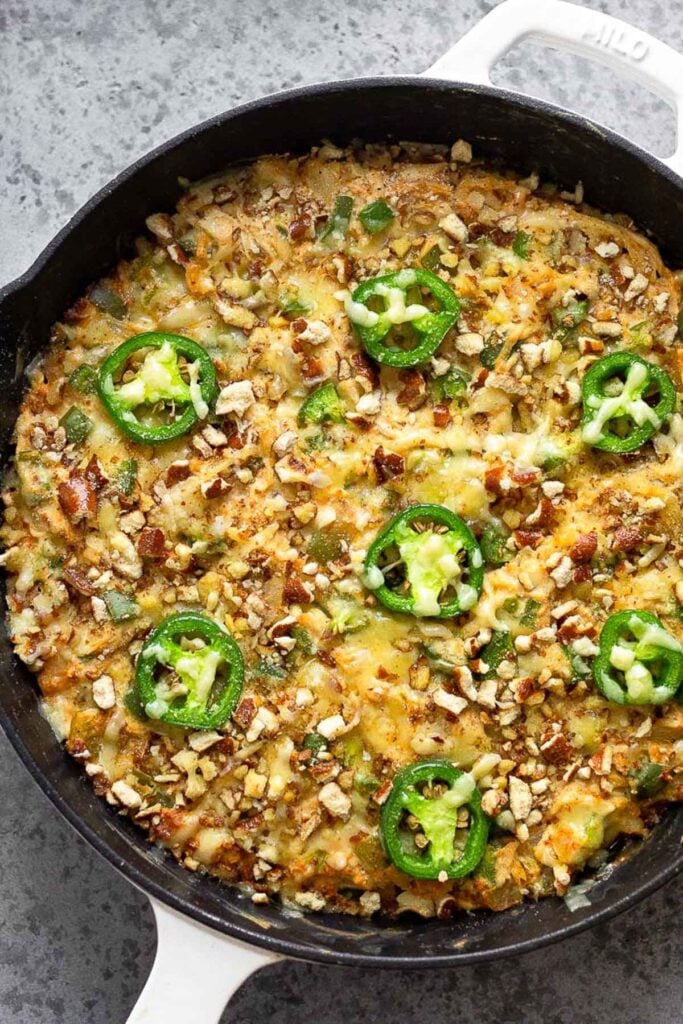 Macro friendly chicken casserole recipe with shredded chicken, veggies, and yogurt that is topped with crushed pretzels, melted cheese, and jalapeños in a white skillet.
