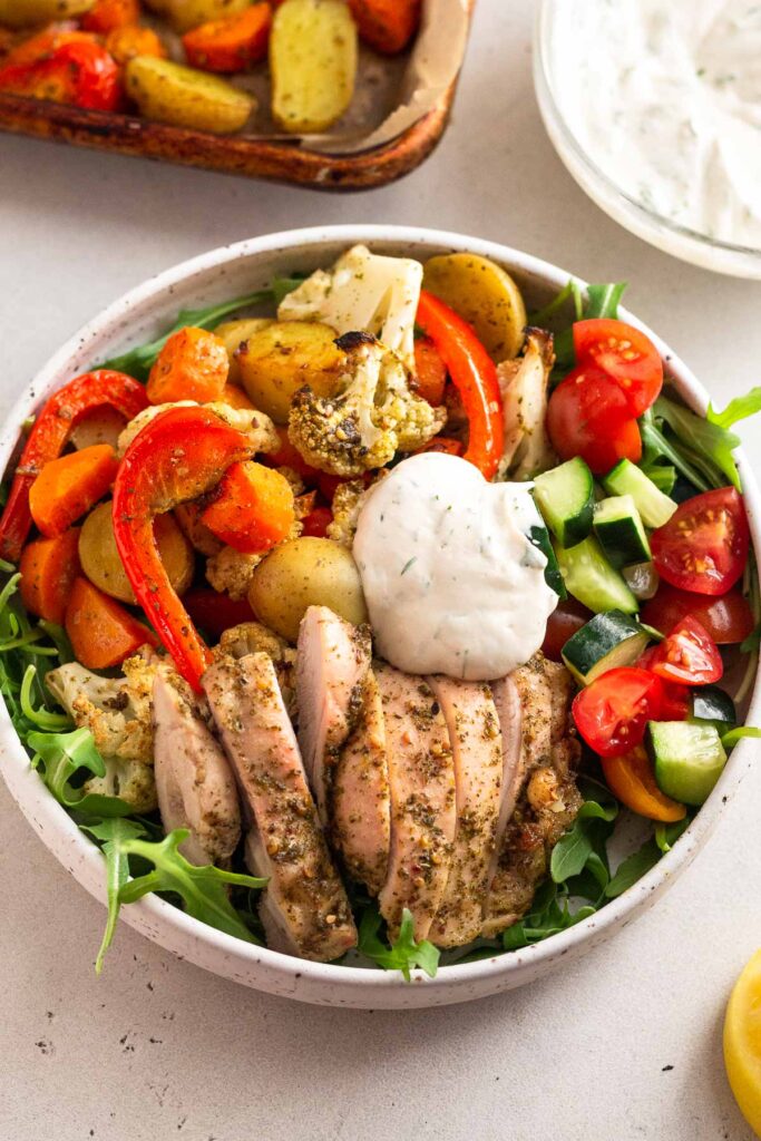 Za’atar chicken bowl with arugula, sliced chicken, roasted potatoes, cauliflower, carrots, peppers, a mix of tomatoes and cucumbers, and a dollop of tahini yogurt sauce. Behind it is a sheet pan and bowl of more sauce.