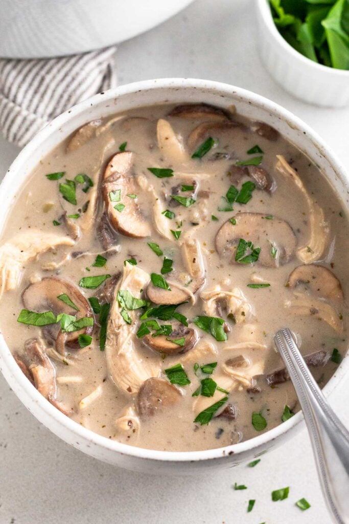 A bowl of creamy chicken mushroom soup topped with fresh parsley. A spoon is in the bowl. Behind it is a ramekin of more herbs and big pot.