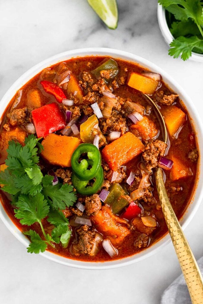 Bowl of chili with butternut squash and peppers. It is topped with cilantro and jalapeños.