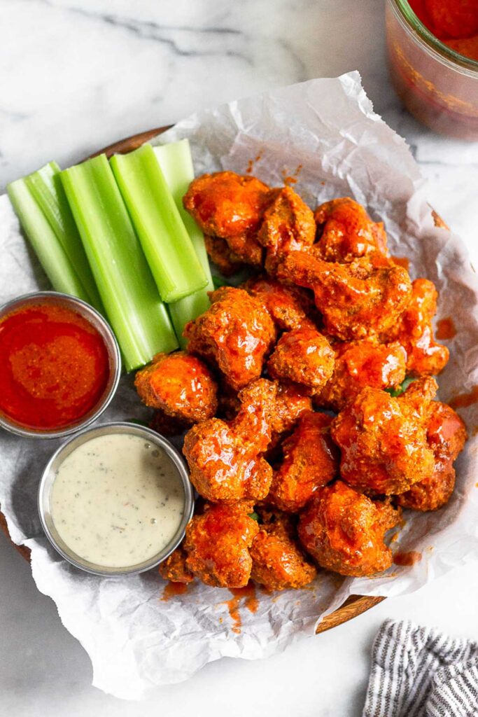 Buffalo cauliflower on a plate with a side of celery, a small dish of buffalo sauce, and a small dish of ranch dressing.