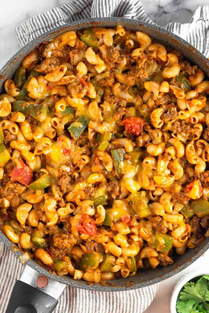 Overhead shot of a large skillet filled with cheesy taco pasta. The skillet is sitting on a kitchen towel next to a small ramekin of cilantro.
