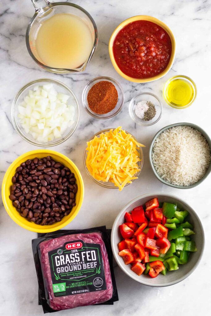 White marble counter top with a bowl of red salsa, a bowl of oil, a bowl of salt and pepper, a bowl of uncooked white rice, a bowl of diced bell peppers, two packages of ground beef on top of each other, a bowl of black beans, a bowl of shredded cheese, a bowl of diced onions, a bowl of spices, and a cup of broth.