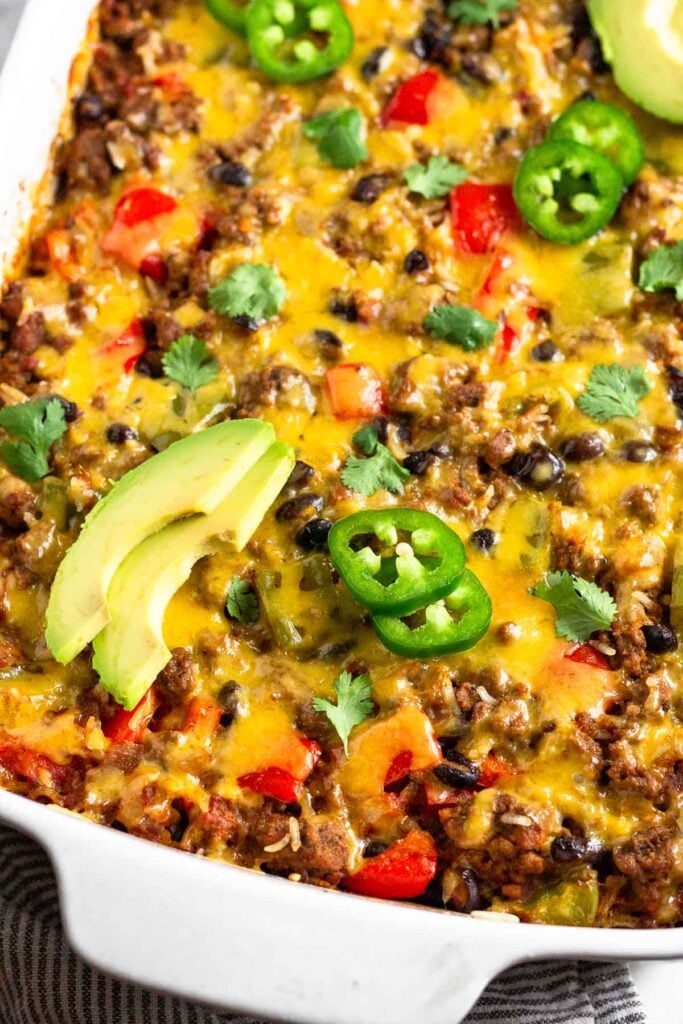 Close up of cheesy beef burrito casserole in a large casserole dish. It is topped with sliced avocado, sliced jalapeño, and cilantro.