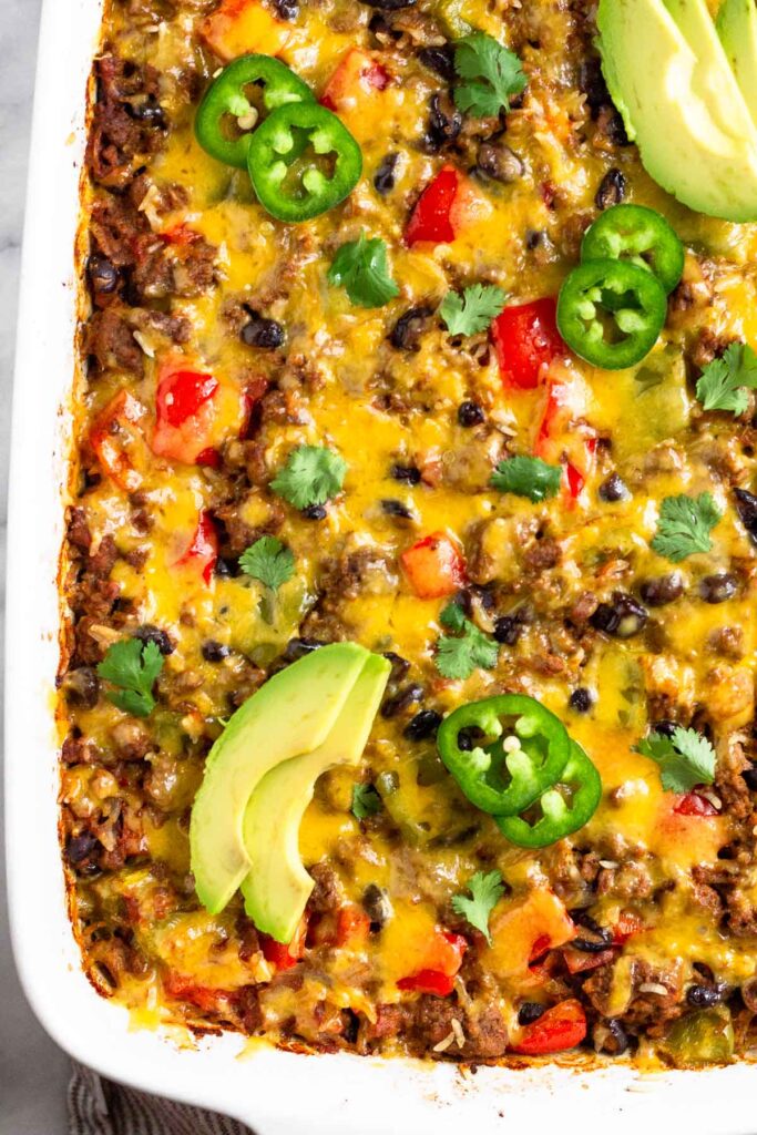 Baked burrito casserole topped with melted cheese in a large casserole dish. It is topped with sliced avocado, sliced jalapeño, and cilantro.