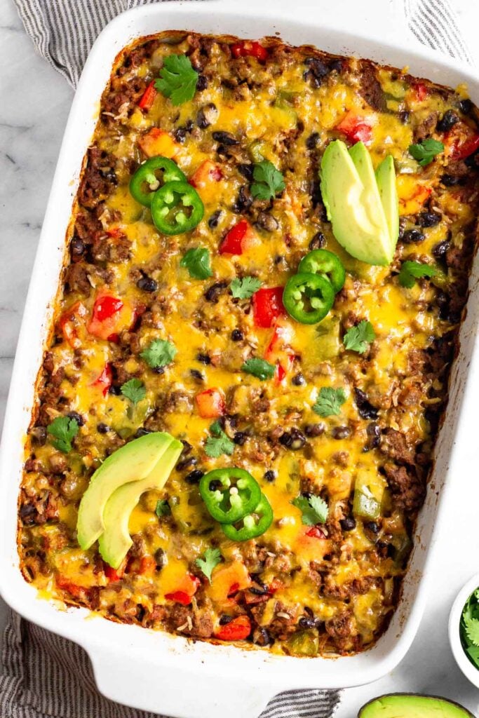 Burrito casserole in a large casserole dish garnished with sliced avocado, sliced jalapeño, and cilantro. Next to it is half an avocado and ramekin of cilantro.