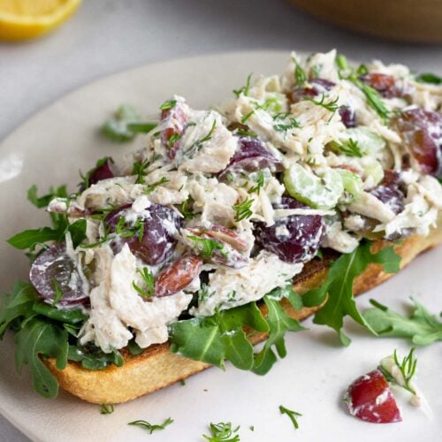 Healthy chicken salad on top of some arugula on a piece of toast on a white plate. Behind it is half a lemon and a bowl of more chicken salad.