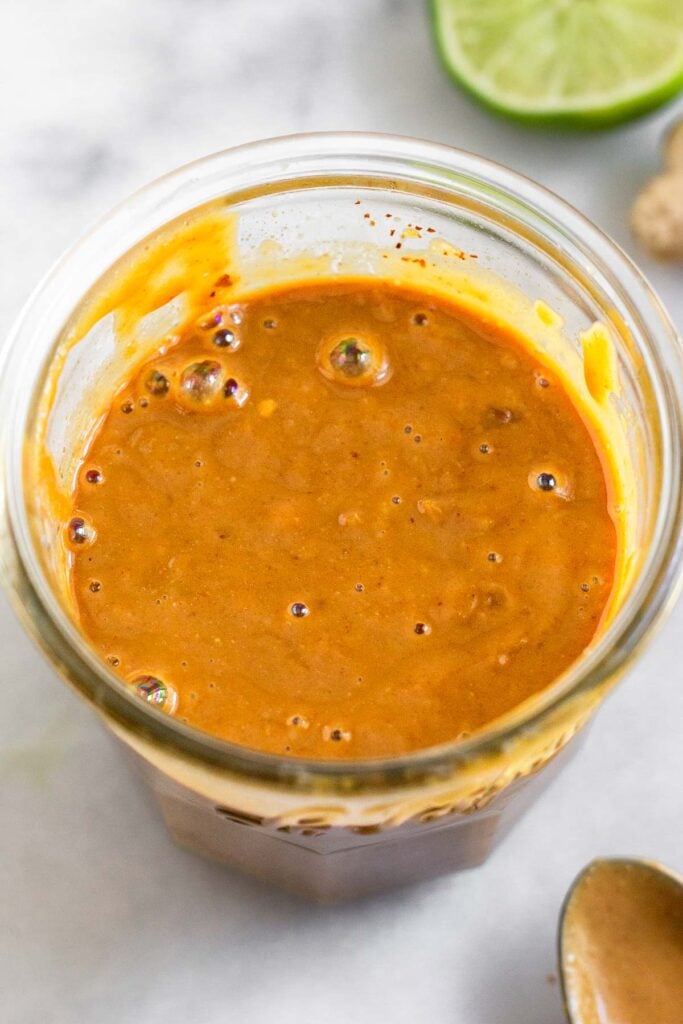 Thai peanut sauce in a medium jar. Around it is a spoon with more sauce on it, fresh ginger, and half a lime.