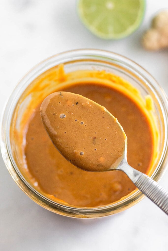 Peanut sauce in a jar with a spoon overtop holding some. Behind the jar is half a lime and fresh ginger.