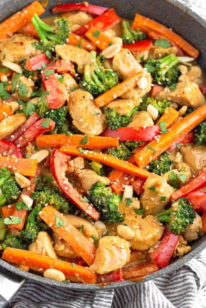A large pan of chicken and veggie stir fry tossed with peanut sauce and topped with cilantro, sesame seeds, and peanuts.