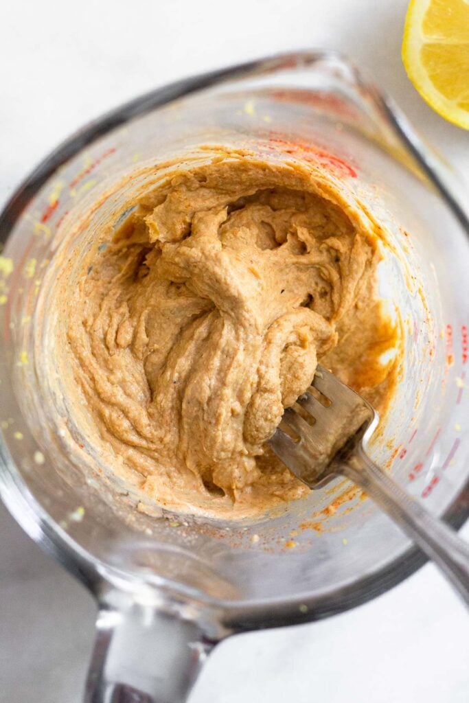 A mixture of tahini a spices in a large measuring sup with a fork in it. Next to the cup is half a lemon.