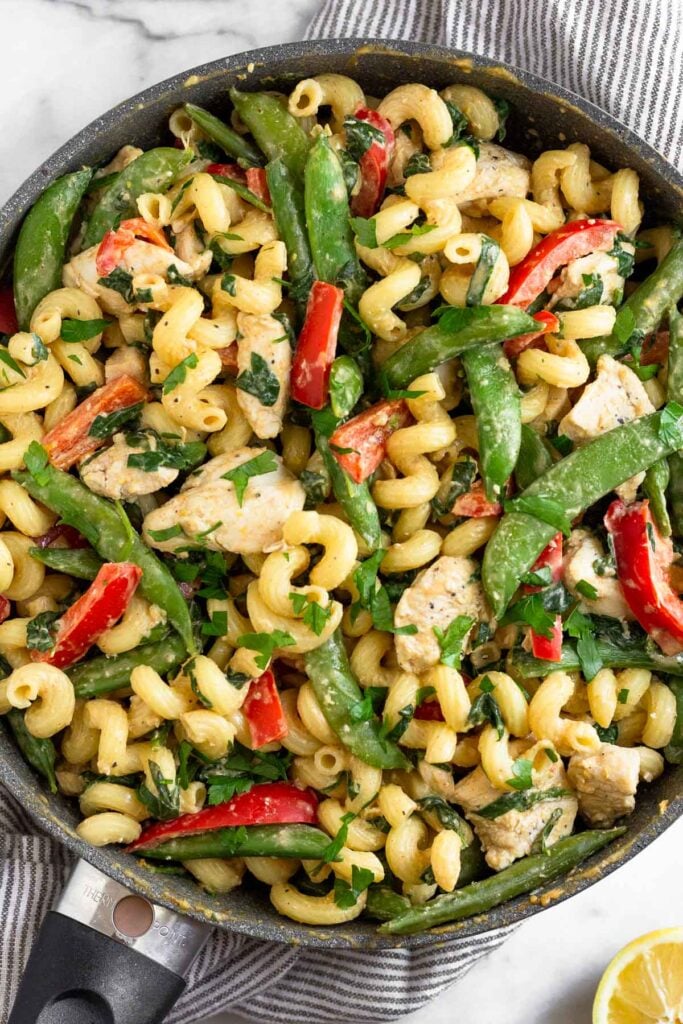 Large skillet filled with chicken pasta and veggies tossed in a tahini sauce. It is garnished with fresh herbs. The pan is on a towel and half a lemon wedge is next to it.