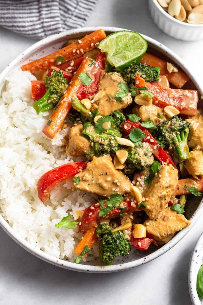 Peanut butter chicken in a bowl with white rice topped with cilantro, peanuts, sesame seeds and a lime wedge. Behind the bowl is a kitchen towel and a small bowl of peanuts.