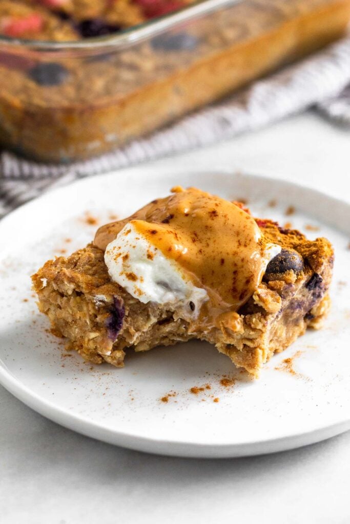 A piece of baked protein oatmeal with a bite taken out of it on a plate topped with yogurt, peanut butter, and cinnamon. Behind it is the pan of the rest of the oatmeal.