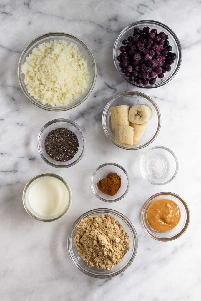 A bowl of frozen blueberries, a bowl of frozen banana pieces, a small bowl of salt, a small bowl of cinnamon, a small bowl of peanut butter, a bowl of vanilla protein powder, a jar of milk, a bowl of chia seeds, and a bowl of frozen cauliflower rice.