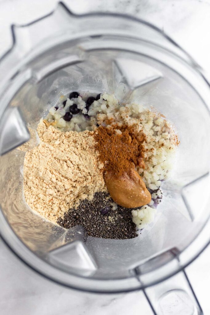 Blender filled with frozen blueberries, frozen cauliflower rice, protein powder, chia seeds, peanut butter, cinnamon, and milk.
