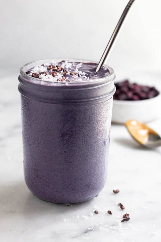 Blueberry hormone balancing smoothie in a jar, topped with coconut shreds and cacao nibs, with a straw. Behind it is a bowl of frozen blueberries and a spoon with peanut butter on it.