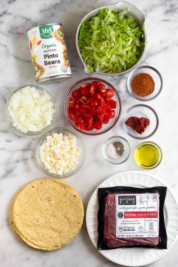 White marble counter with a bowl of shredded lettuce, a bowl of taco seasoning, a bowl of tomato paste, a bowl of olive oil, a plate with a package of ground beef on it, a stack of corn tortillas, a bowl of salt and pepper, a bowl of crumbled cheese, a bowl of diced tomatoes, a bowl of diced onions, and a can of refried beans.