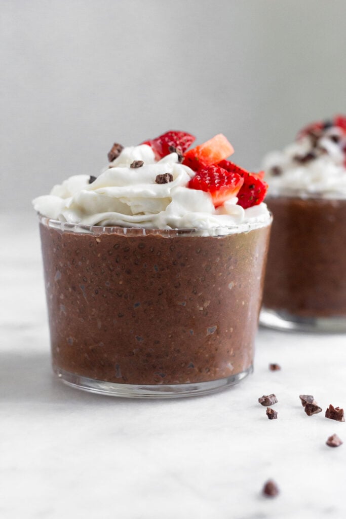 Chocolate chia pudding in a small glass topped with whipped cream, strawberries, and cacao nibs. Behind it is a similar glass and around them is cacao nibs.